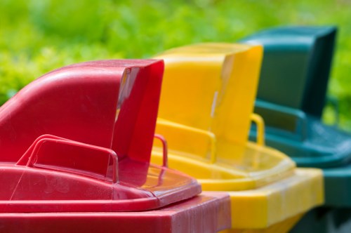 A row of four colourful plastic bins positioned outdoors, likely on a driveway or pavement near a green grassy area. The first bin in the foreground is red with a smooth, slightly shiny surface, followed by a yellow bin with a similar finish, then a green bin, and finally a blue bin partially visible behind the others. Each bin has a raised lid and integrated handles on the sides. The bins appear to be used for waste or recycling collection, designed for outdoor rubbish removal services such as those provided by Commercial Waste Removal Hatch End. The background includes a blurred view of natural greenery, suggesting a residential or suburban setting in the Hatch End area, within the local postcode vicinity. The lighting is natural daylight, providing clear visibility of the textured surfaces and vivid colours of the bins, emphasizing their durable plastic construction suited for waste disposal tasks.