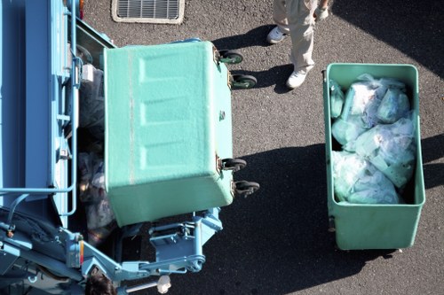 An overhead view of a green wheelie bin filled with transparent plastic bags containing waste, positioned next to a larger green recycling container on a paved surface. The recycling bin is partly filled and open, revealing the waste inside. A person wearing beige trousers and dark shoes stands nearby, indicating an active rubbish collection or disposal scene. The curbside setting suggests a typical residential or commercial waste collection area, with part of a blue waste disposal vehicle visible on the left side of the image. The environment is outdoor, with the dark asphalt surface and natural daylight casting shadows, illustrating a typical rubbish removal operation in Hatch End, London. The image visually supports services offered by Commercial Waste Removal Hatch End for rubbish disposal.