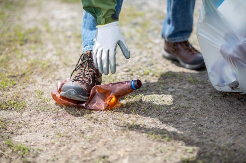A person wearing white gloves and jeans is seen placing a brown glass bottle onto the ground, with an outdoor setting featuring a dirt path and green grass nearby. In the background, another individual dressed in blue denim and brown footwear is holding a transparent plastic rubbish bag that appears to contain additional waste. The scene suggests a cleanup or rubbish removal activity, with the focus on proper disposal of litter such as glass bottles, aligned with services offered by Commercial Waste Removal Hatch End for waste management and rubbish collection in the local area. The environment is well-lit, indicating daytime, and the ground surface shows natural soil and grass, contributing to the impression of an outdoor residential or communal space in Hatch End, Middlesex, covered under postcode HA5. This image exemplifies responsible waste disposal practices, suitable for discussing local rubbish removal services.