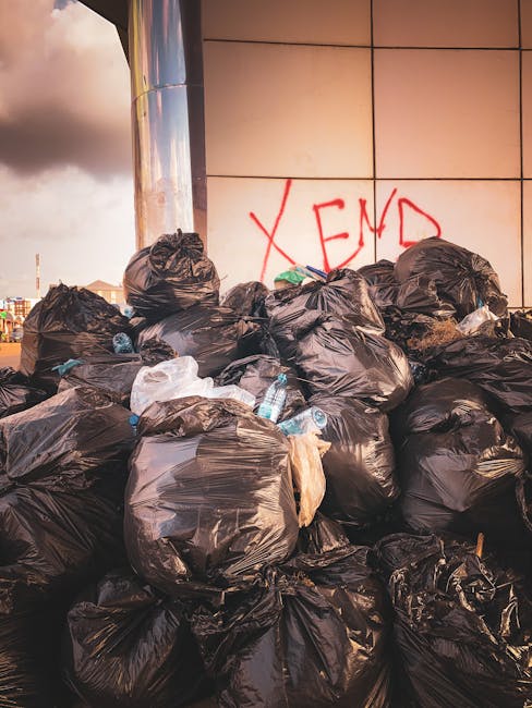 A large pile of black rubbish bags filled with waste is stacked against a tiled wall outdoors. Some bags appear to be partially torn or leaning, with plastic bottles and miscellaneous rubbish visible 