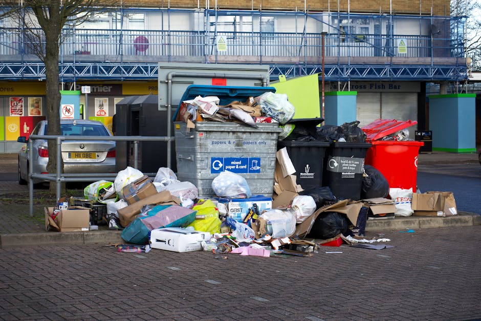 A pile of rubbish accumulated on a concrete driveway surrounded by a low brick wall and a garden area. The waste includes various materials such as large black plastic bags filled with debris, cardboa
