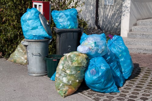 A collection of black, grey, and green rubbish bins and several large blue and clear plastic bags filled with waste are arranged on a paved driveway near a garden area, with some bags leaning against or stacked beside the bins. The plastic bags contain a mixture of household waste and recyclable materials, visible through the semi-transparent plastic. The scene is outdoors, adjacent to a residential property, with a flower bed and greenery in the background, as well as a section of grey concrete steps leading to a building entrance. The lighting indicates daylight, possibly overcast, creating a neutral and clear view suitable for a rubbish removal service website such as Commercial Waste Removal Hatch End, serving local businesses and residents in the Hatch End area, captured in a straightforward and factual style to support accessibility and local SEO relevance. The image emphasizes waste collection and disposal suitable for rubbish removal services in the area postcode detailed in the page, with a focus on organized waste accumulation ready for collection.